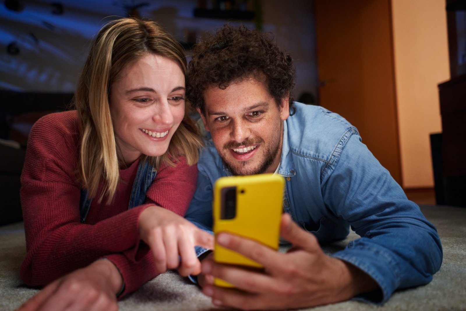 Happy nice Caucasian couple using phone smiling lying on carpet in living room at home at night.